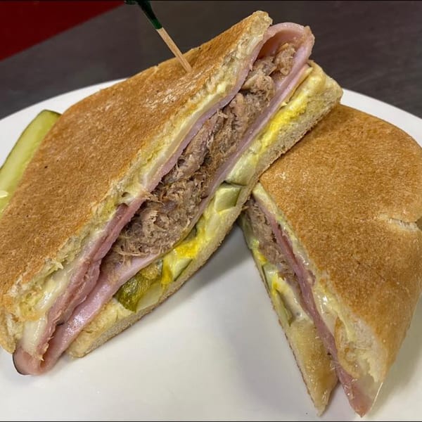 A Cuban sandwich from Old Town Red Rooster Cafe, cut in half, with a pickle spear on a white plate.