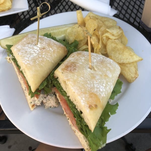 Two ciabatta sandwiches with lettuce, tomato, and chicken salad, served with chips and a pickle.
