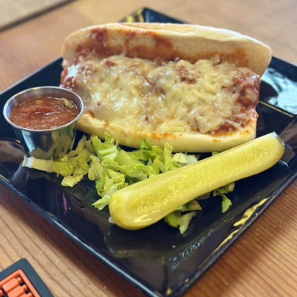 A meatball sub with melted cheese, a side of marinara sauce, lettuce, and a pickle spear.