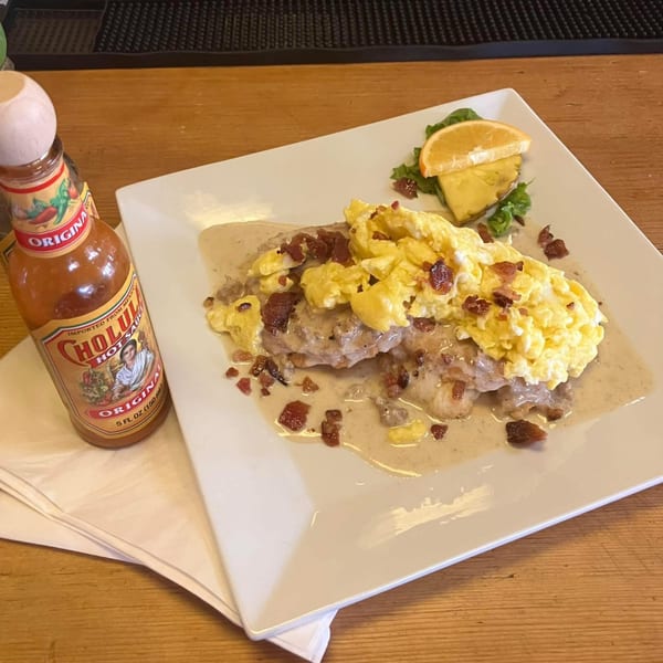 A breakfast plate with biscuits and gravy, scrambled eggs, bacon bits, and a side of fruit.