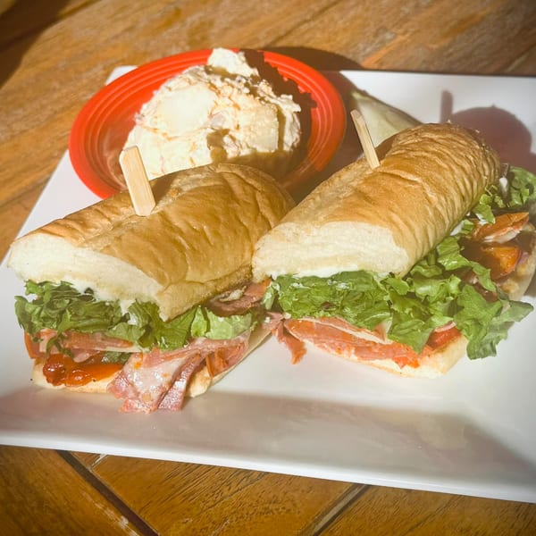 A large deli sandwich, cut in half, with lettuce, meat, and sauce, served with potato salad.